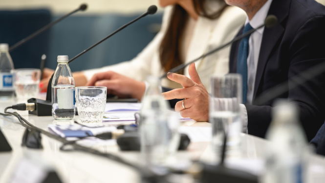 Tuvplānā redzama galda virsma uz kuras uzlikti dokumenti, galda mikrofoni un ūdens pudeles ar glāzēm, tāluma redzami cilvēku silueti