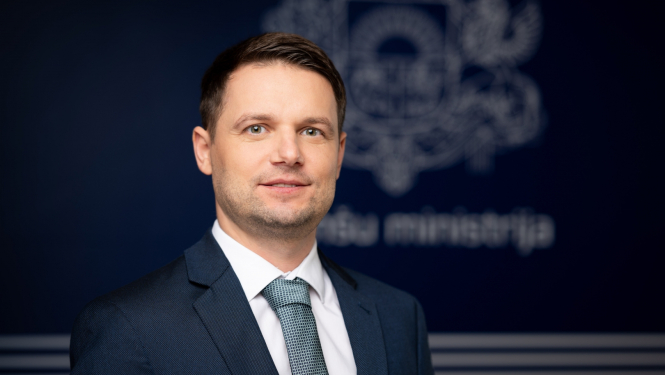 Vīrieša zilā uzvalkā ar kaklasaiti portrets uz zila fona ar Finanšu ministrijas ģērboni