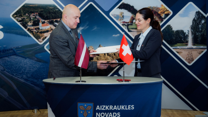 Vīrietis un sieviete apmainās ar dokumentu mapēm, priekšplānā uz galda - Latvijas un Šveices karodziņi