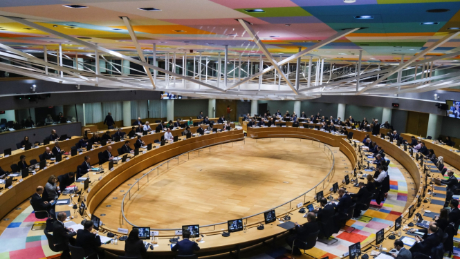 Daudz cilvēku sēž milzīgā zālē pie ovāla gada