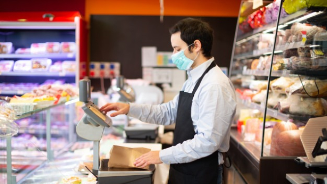 pārdevējs sejas maskā sver preci veikalā aiz letes