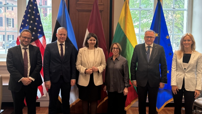 Lielu logu un dažādu valtu karogu fonā rindā stāv vīrieši un sievietes, priekšplānā redzama galda daļa