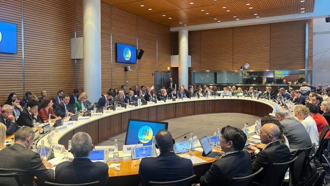 Pie ovāla galda sēž vīrieši un sievietes, un piedalās diskusijā