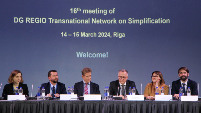 Pie galda rindā sēž sievietes un vīrieši