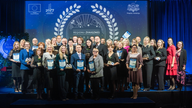 Uz skatuves stāv grupa cilvēku ar diplomiem rokās