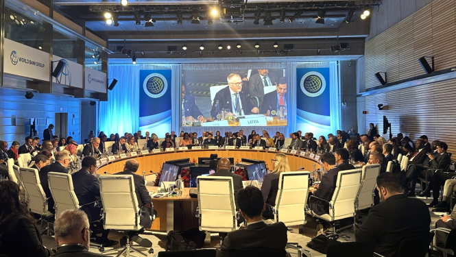 Lielā zālē pie ovāla galda sēž daudz vīrieši un sievietes un diskutē