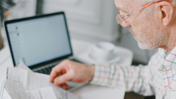 Persona ar brillēm pie darba galda pārskata papīra dokumentus, fonā atvērts klēpjdators