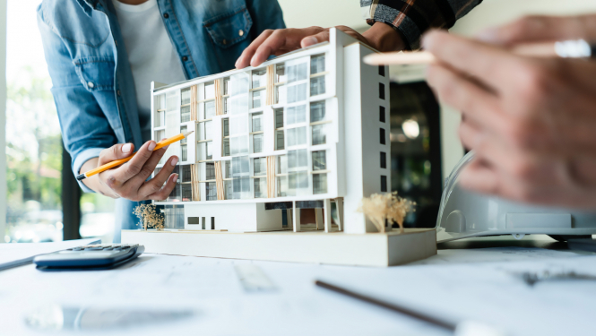 Tuvplānā redzams daudzstāvu ēkas modelītis un cilvēka rokas