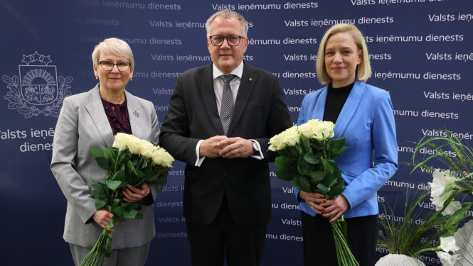 Sieviete gaišā kostīmā tur rokās baltu rožu pušķi, vīrietis tumšā uzvalkā un sieviete zilā žaketē tur rokās baltu rožu pušķi