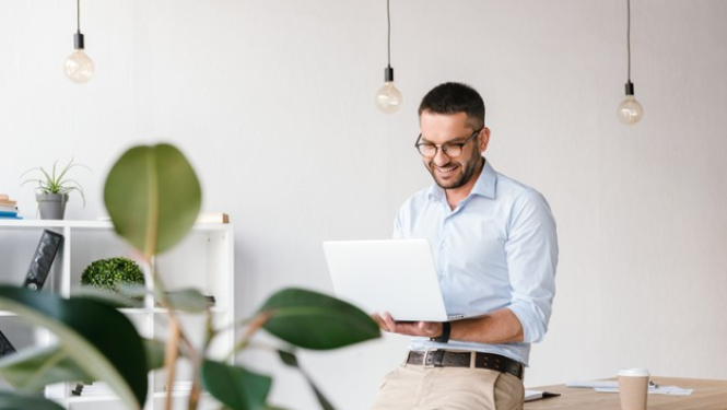 Smaidīgs vīrietis skatās datorā, kuru tur rokās