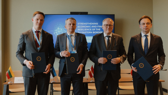 Četri vīrieši stāv rindā, tur rokās mapes ar dokumentiem, aiz viņu mugurām stāv gals ar krēsliem, pie sienas TV ekrāns