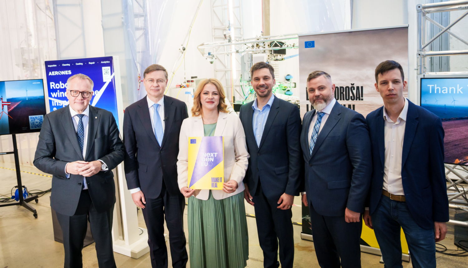 Industriālā vidē rindā sastājušies pieci vīrieši un viena sieviete, skatās kamerā un smaida. Sievietei rokās ir dokumentu mape