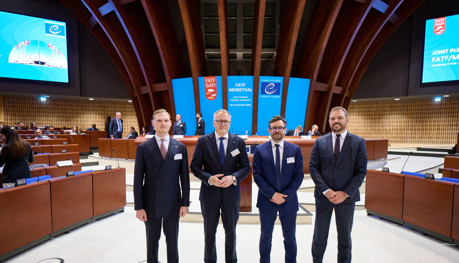 Attēlā redzami četri vīrieši uzvalkos, kuri pozē oficiālā vidē – konferenču zālē