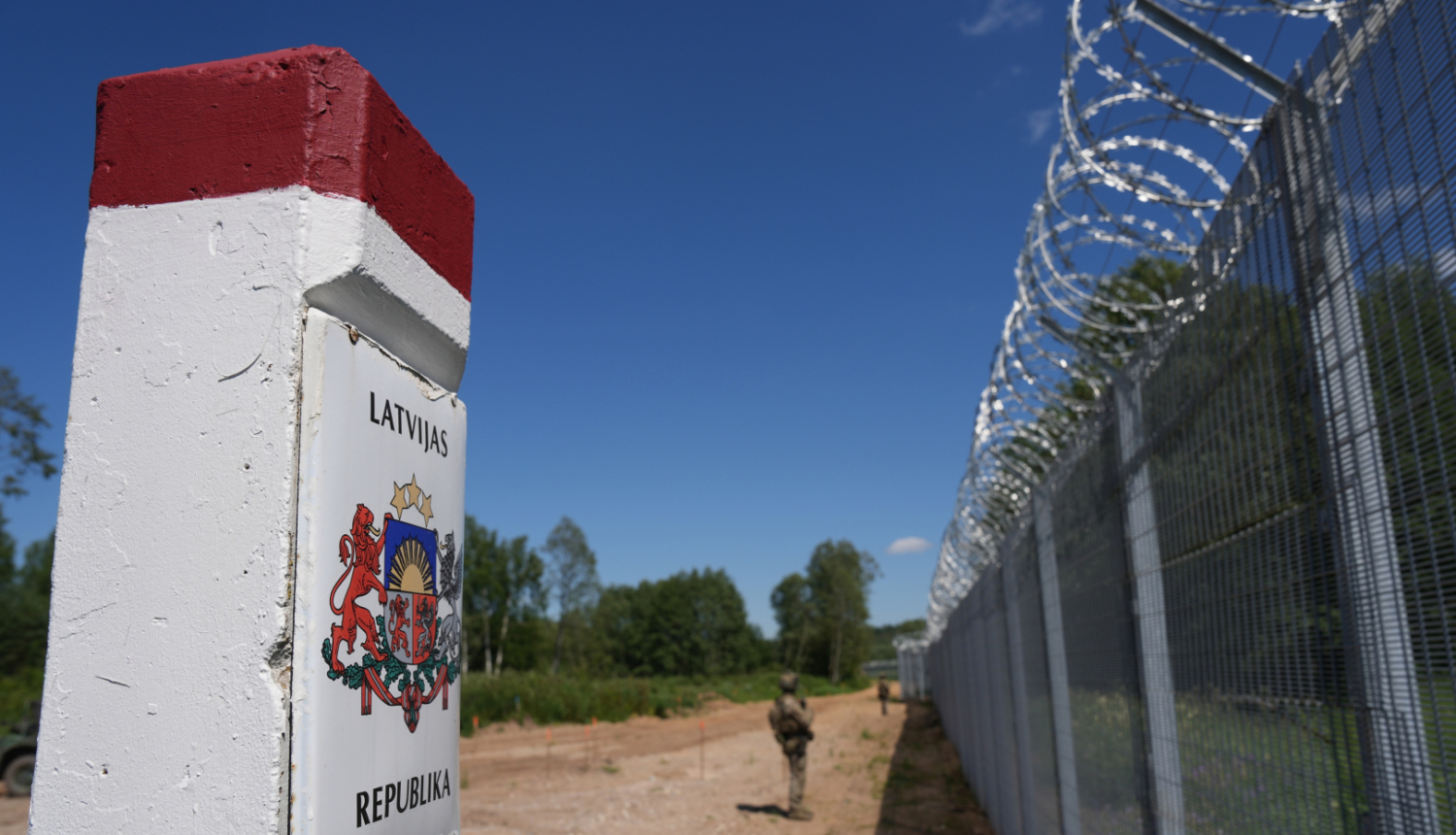 Attēlā tuvplānā redzams robežstabiņš ar Latvijas ģerboni, līdzās augstam žogam ar dzeloņstieplēm