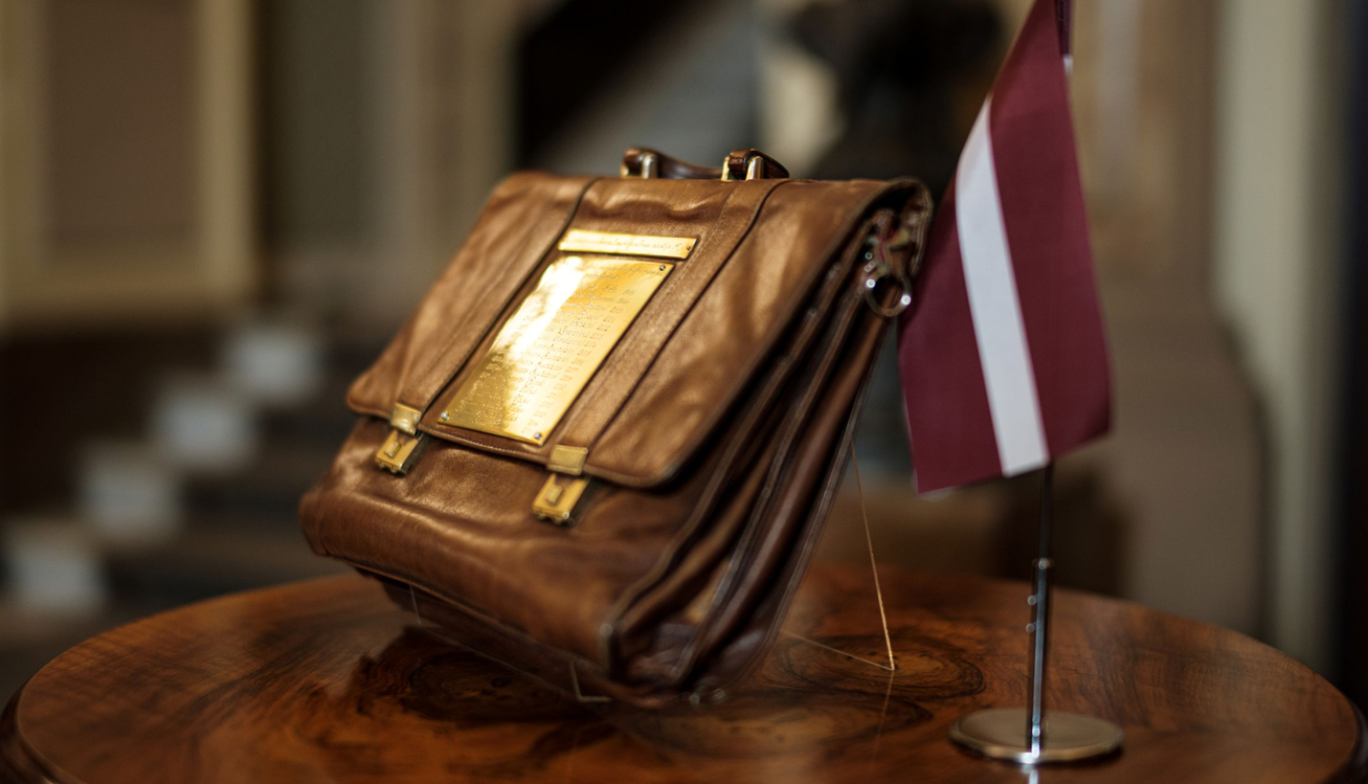 Attēlā redzams tradicionālais budžeta portfelis statīvā novietots uz apaļa galdiņa, līdzās stāv Latvijas karodziņš