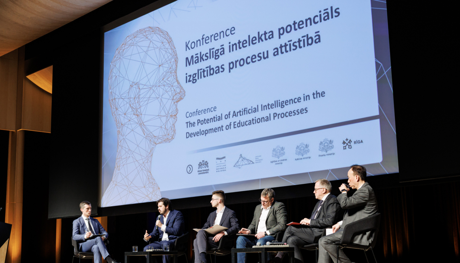 Attēlā redzami seši vīrieši, kuri sēž krēslos uz skatuves un savstarpēji diskutē, fonā milzīgs projektora ekrāns