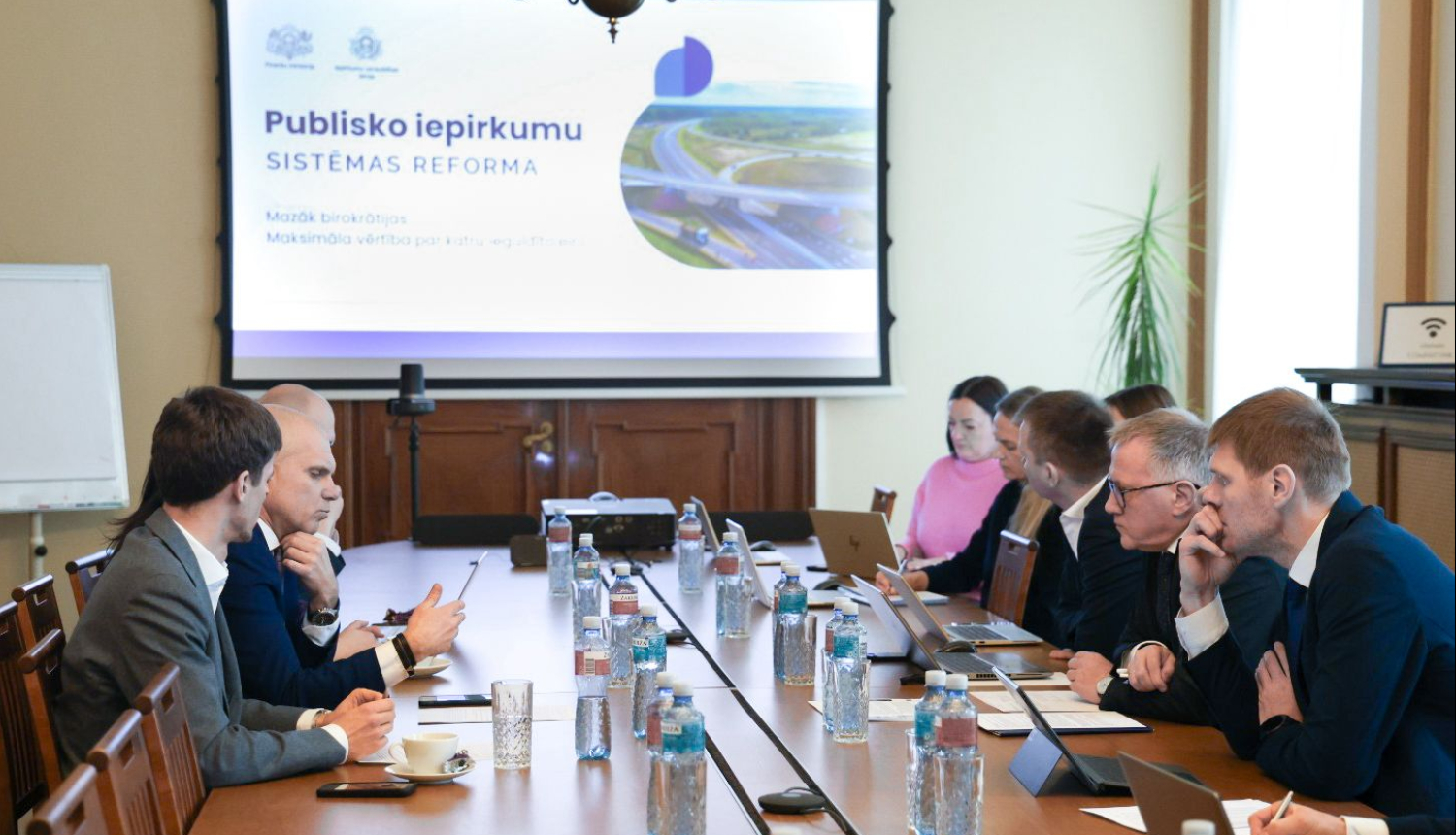 Pie gara sēžu galda abās pusēs sēž vīrieši un sievietes, savstarpēji diskutē, attēla centrā projektora ekrāns, uz kura redzama prezentācija 