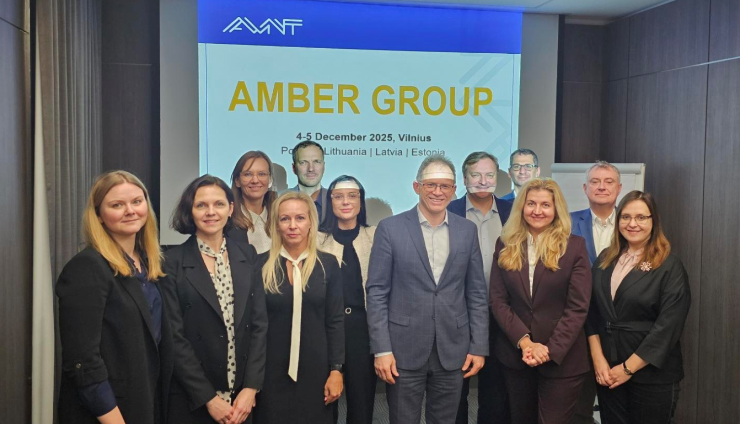 Attēlā redzama grupa cilvēku, kuri stāv vairākās rindās, viņiem aiz muguras redzams projektora eksrāns