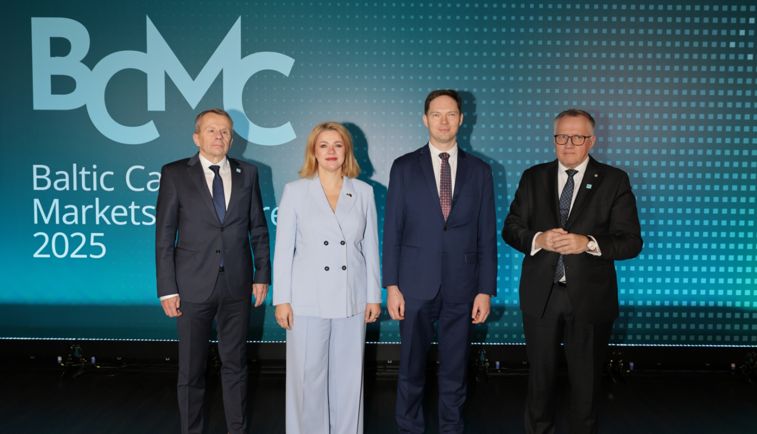 Attēlā redzami trīs kungi uzvalkos un sieviete gaiši zilā bikškostīmā