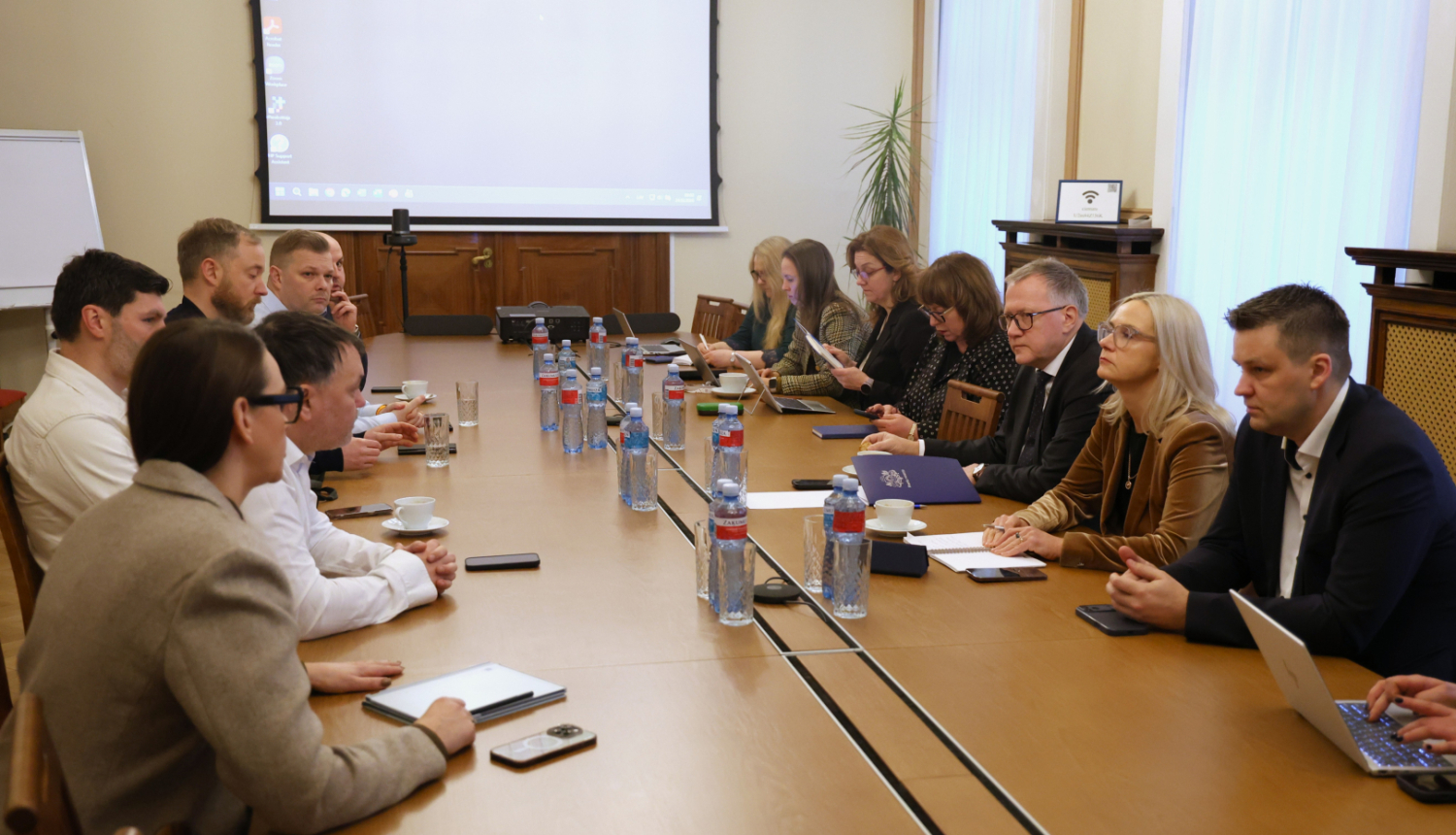 Pie garena galda sēž divas cilvēku grupas, kas apspriež darba jautājumus. Uz galda redzami klēpjdatori, dokumenti, telefoni un ūdens pudeles, fonā projekcijas ekrāns