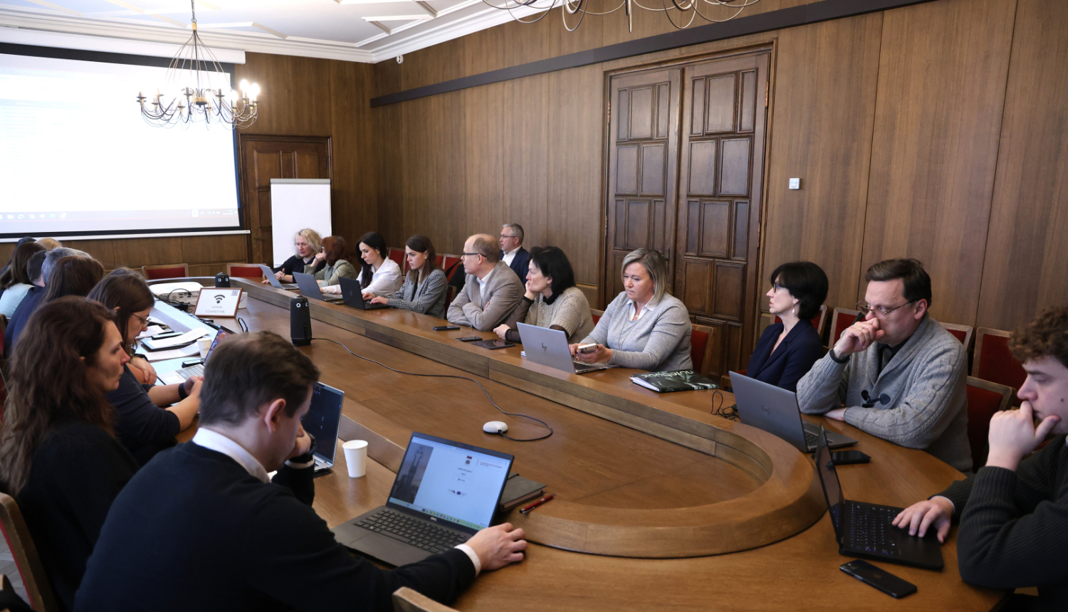 Pie garena sanāksmju galda sēž sievietes un vīrieši, un diskutē, uz galda stāv portatīvie datori un dokumenti