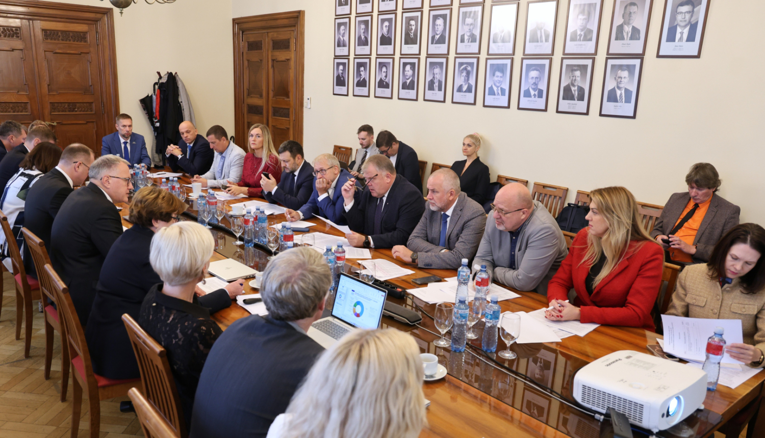 Attēlā garš galds ap kuru no visām pusēm sēž vīrieši un sievietes un savstarpēji sarunājas, uz galda stāv dokumenti, portatīvie datori, ūdens pudeles un glāzes