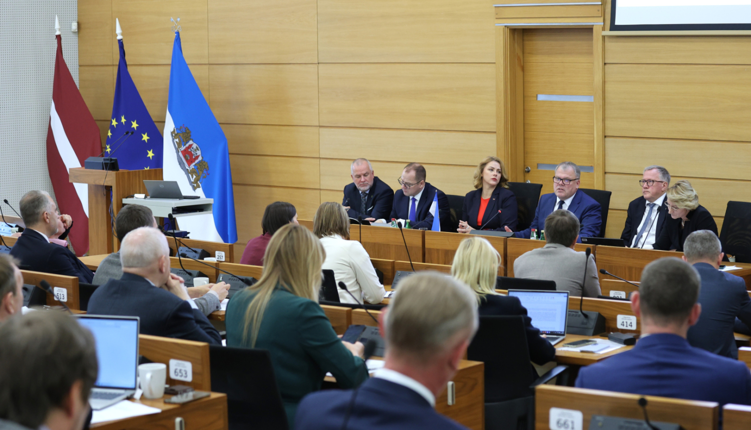 Pie gara galda auditorijas priekšā sēž seši cilvēki, kuri diskutē ar auditoriju, telpas malā stāv Latvijas ES un Rīga domes karogi