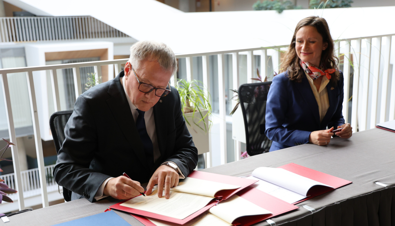 Attēlā vīrietis paraksta uz galda stāvošus dokumentus, blakus sēdošā sieviete skatās uz viņu