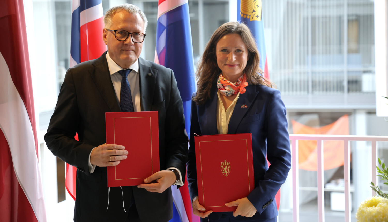 Vīrietis un sieviete uz karogu fona stāv līdzās un smaida, rokās turot dokumentu mapes