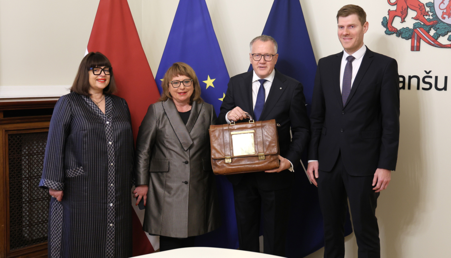 Attēlā uz Latvijas, ES un Nato karogu fona stāv četri cilvēki un smaida, divi no tiem rokās tur portfeli