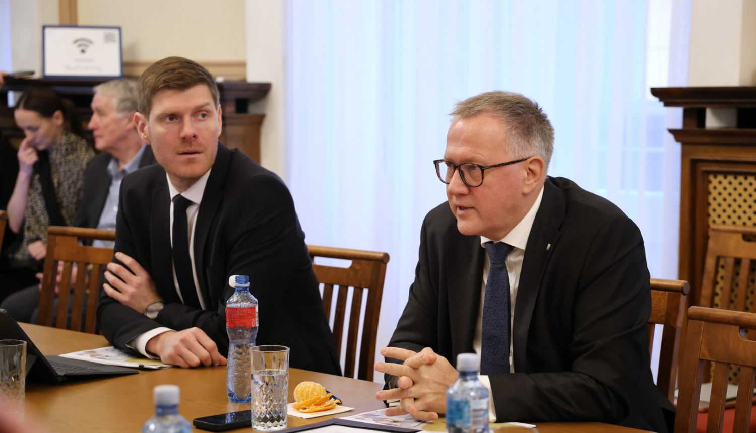 Attēlā pie galda sēžam redzami divi vīrieši uzvalkos, tālumā ir redzami vēl cilvēki sēžam, uz galda stāv dokumenti un ūdens pudeles