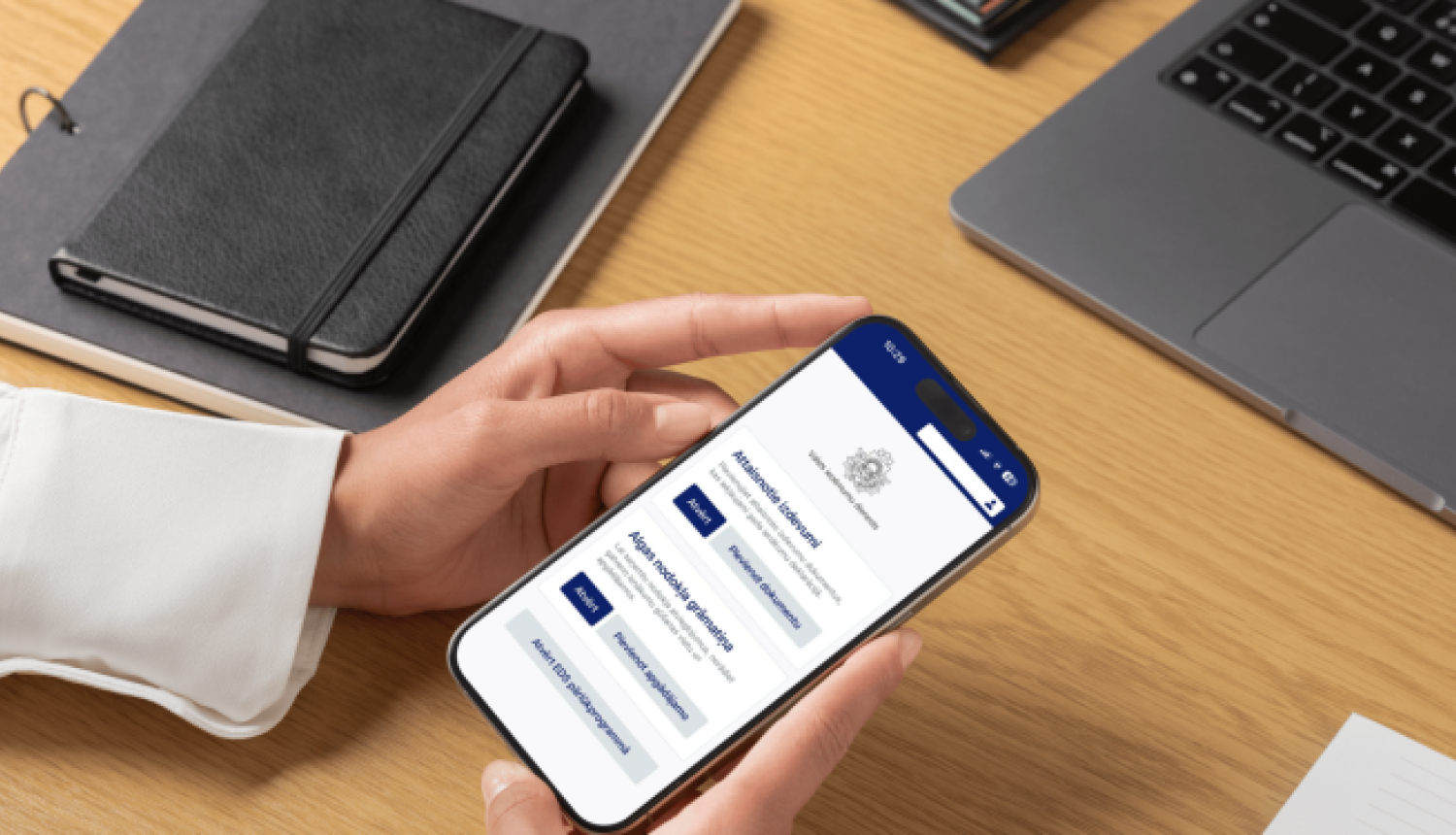 Virs galda virsmas sieviete tur rokās mobilo tālruni, kurā redzama jaunā VID vietne