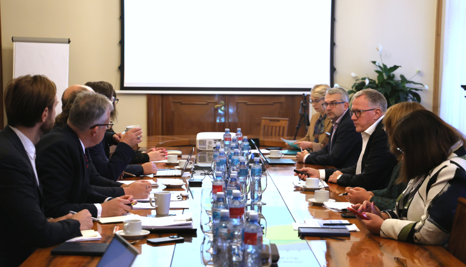 Pie gara galda no abām pusēm sēž vīrieši un sievietes, savstarpēji sarunājas, pie sienas redzams ekrāns, uz kura rāda prezentāciju