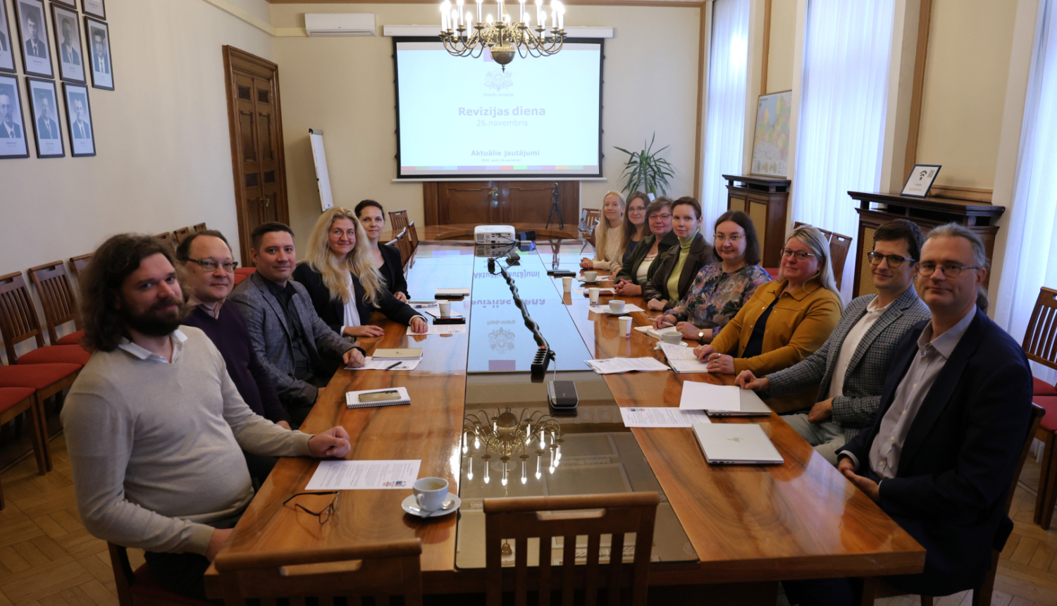 Attēlā pie gara galda abpus galdam sēž vīrieši un sievietes un smaidot skatās kamerā, tālumā pie sienas ir redzams projektora ekrāns