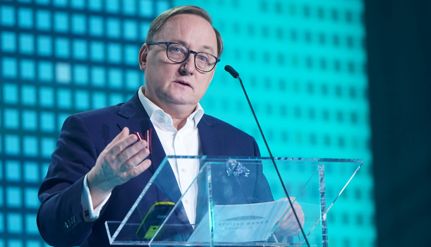 Attēlā tuvplānā redzams vīrietis, kurš stāv pie caurspīdīgas tribīnes un žestikulējot runā, viņas priekšā ir mikrofons