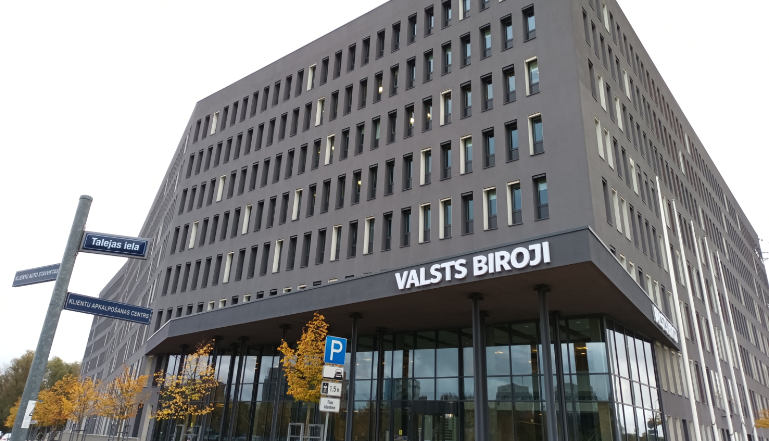 Attēlā redzama pelēka daudzstāvu biroju ēka ar stiklotu ieejas fasādi, uz tās redzams uzraksts VALSTS BIROJI