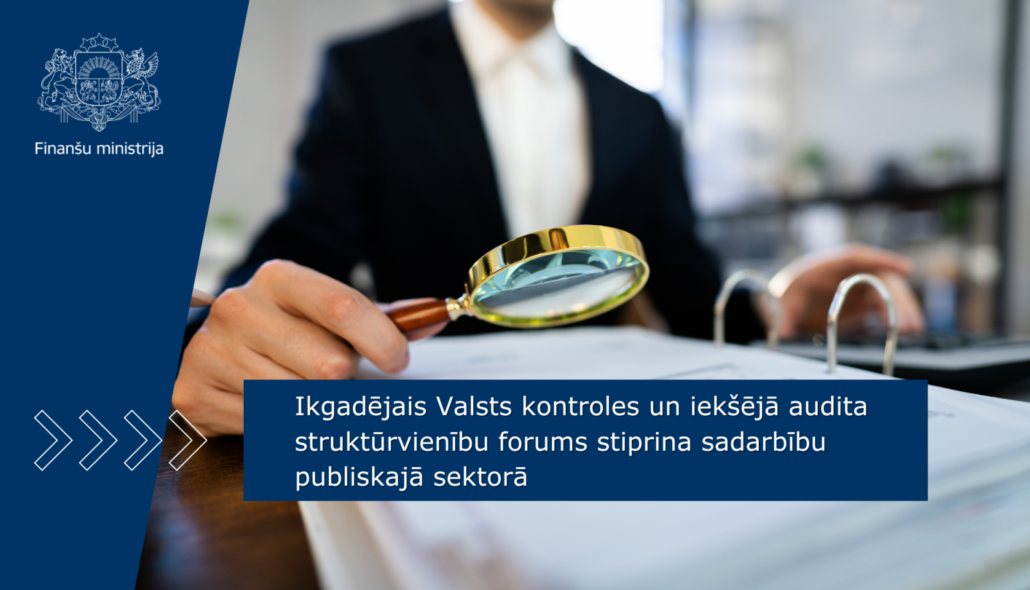 Attēlā redzams cilvēks melnā kostīmā un baltā kreklā sēžam pie galda, rokās tur lupu, kas novietota virs dokumentu kaudzes