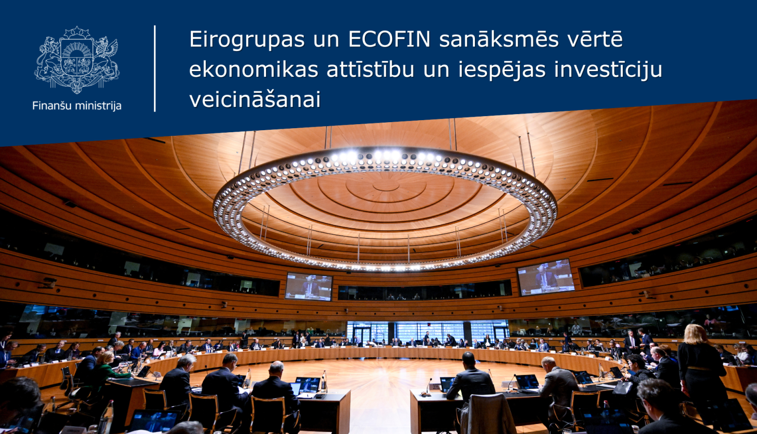 Attēlā redzama plaša apaļa zāle, kurā aplī vairākās rindās izkārtoti galdi, pie kuriem sēž cilvēki, pie griestiem redzama liela apļveida lampa