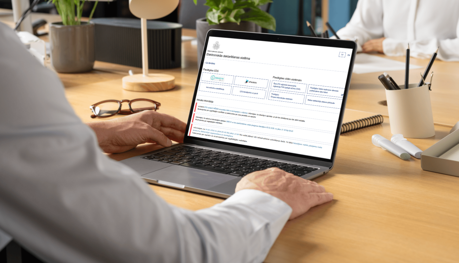 Persona birojā pie galda strādā ar portatīvo datoru, kura ekrānā atvērta Elektroniskās deklarēšanas sistēmas tīmekļa vietne