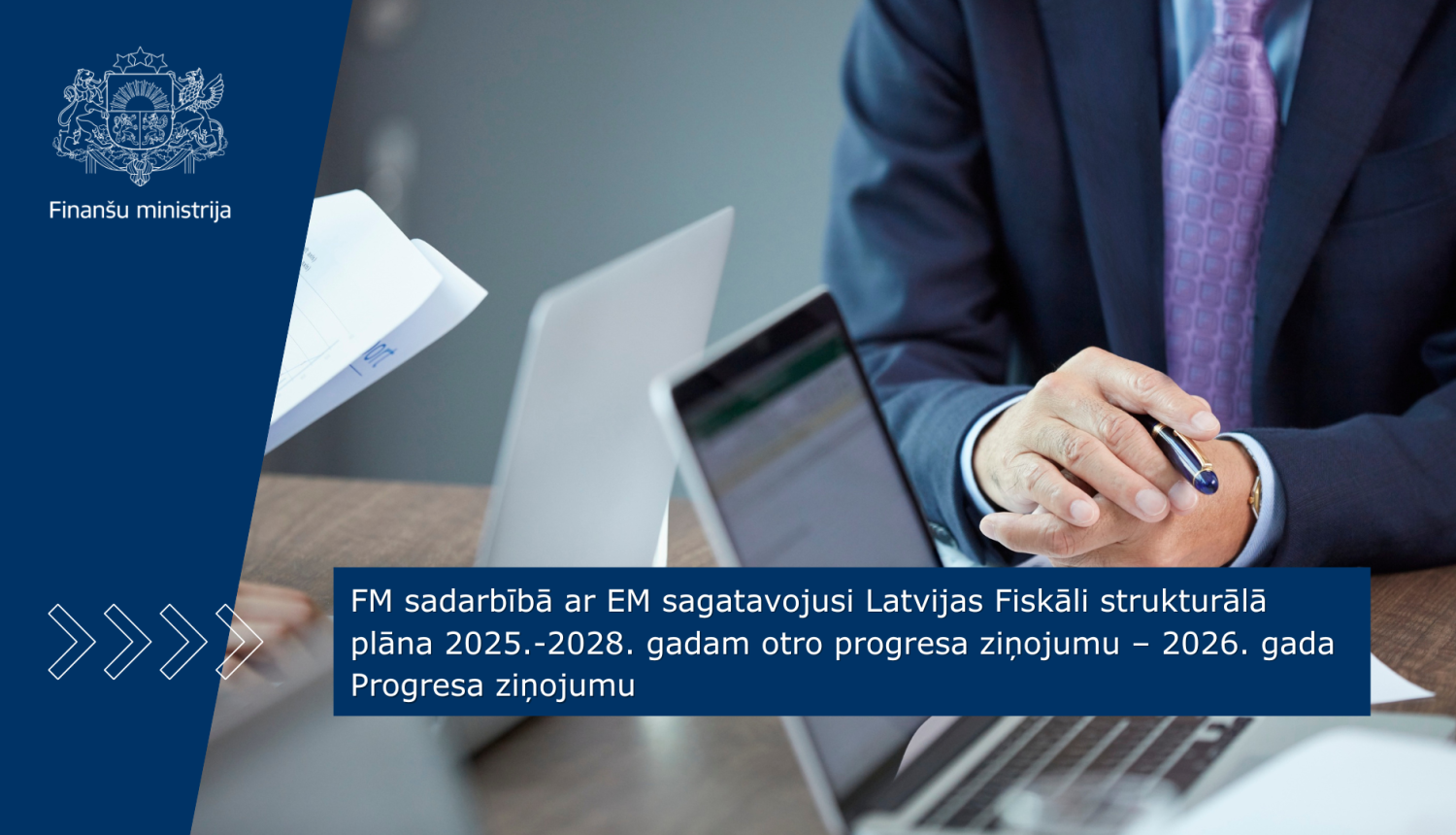 Pie galda sēž vīrietis zilā uzvalkā un diskutē, uz galda stāv vairāki portatīvie datori 