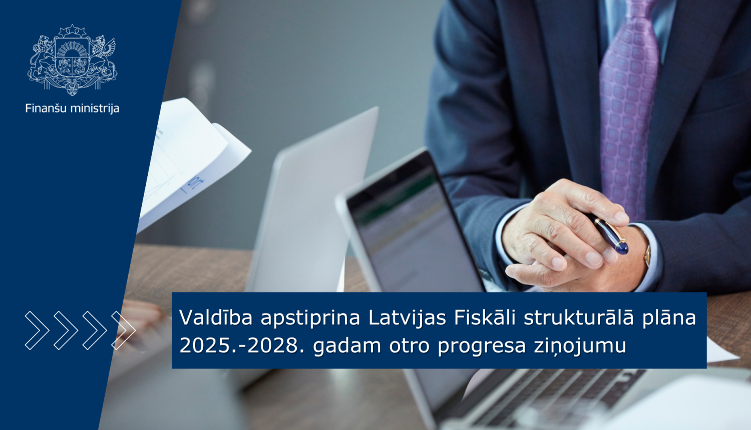 Vīrietis uzvalkā pie galda sēž un strādā ar dokumentiem un portatīvo datoru