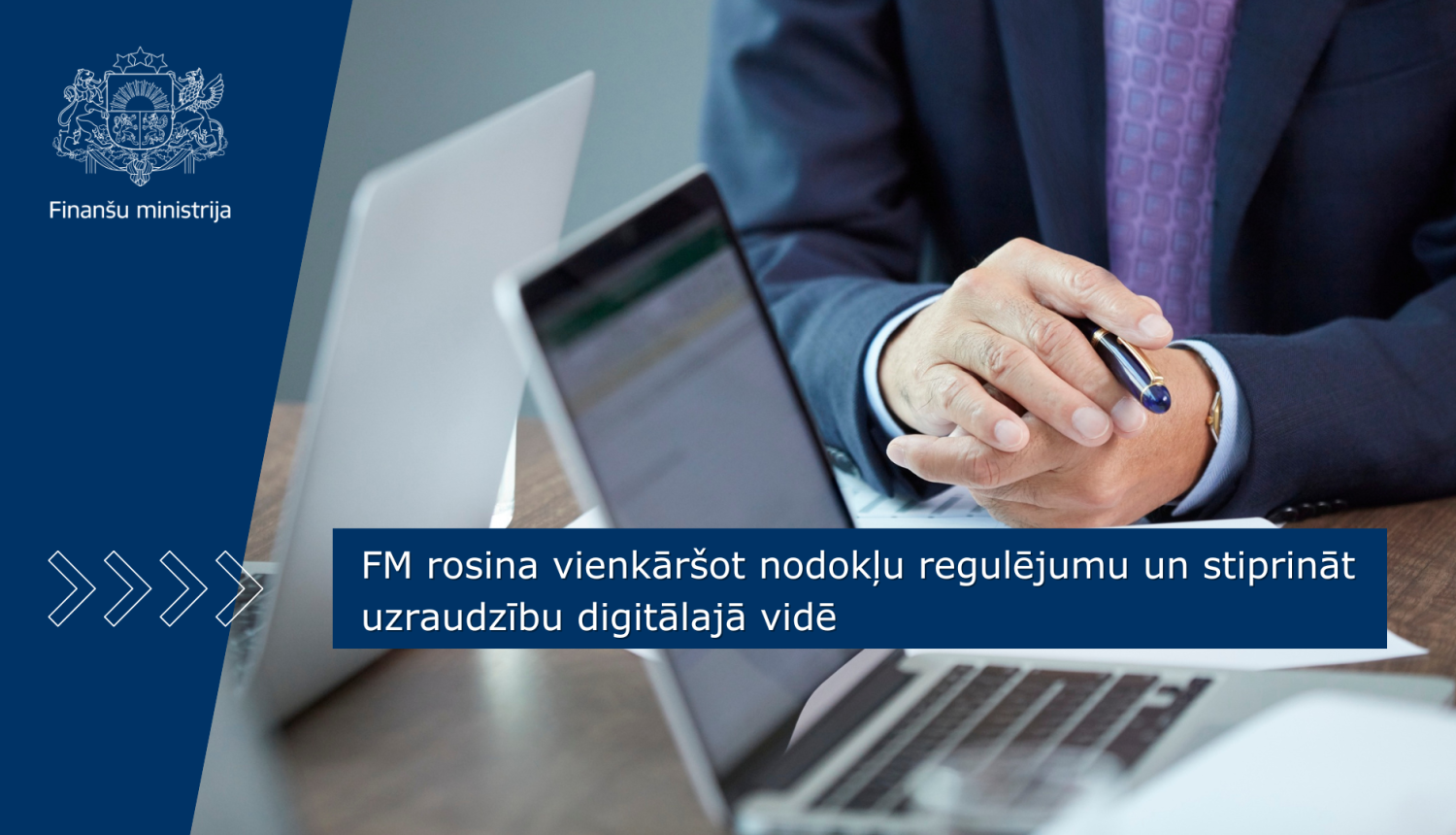 Vīrietis sēž pie galda, tur rokās pildspalvu un norāda uz uz galda esošajiem dokumentiem un portatīvo datoru