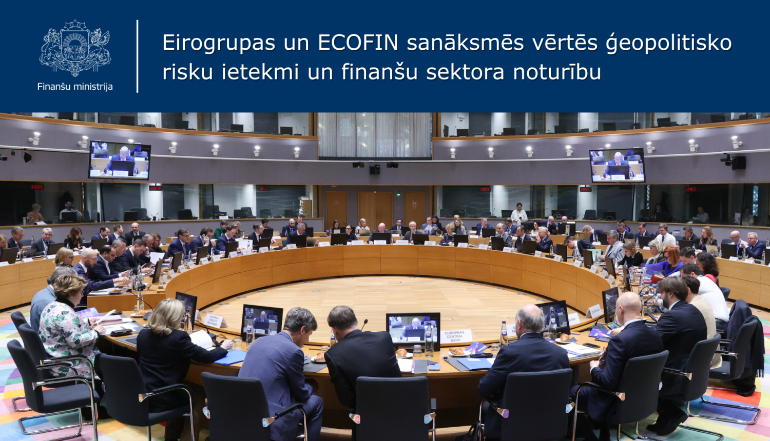 Plašā konferenču zālē ar balkonu pie pa vidu novietota apaļa konferenču galda sēž sievietes un vīrieši 