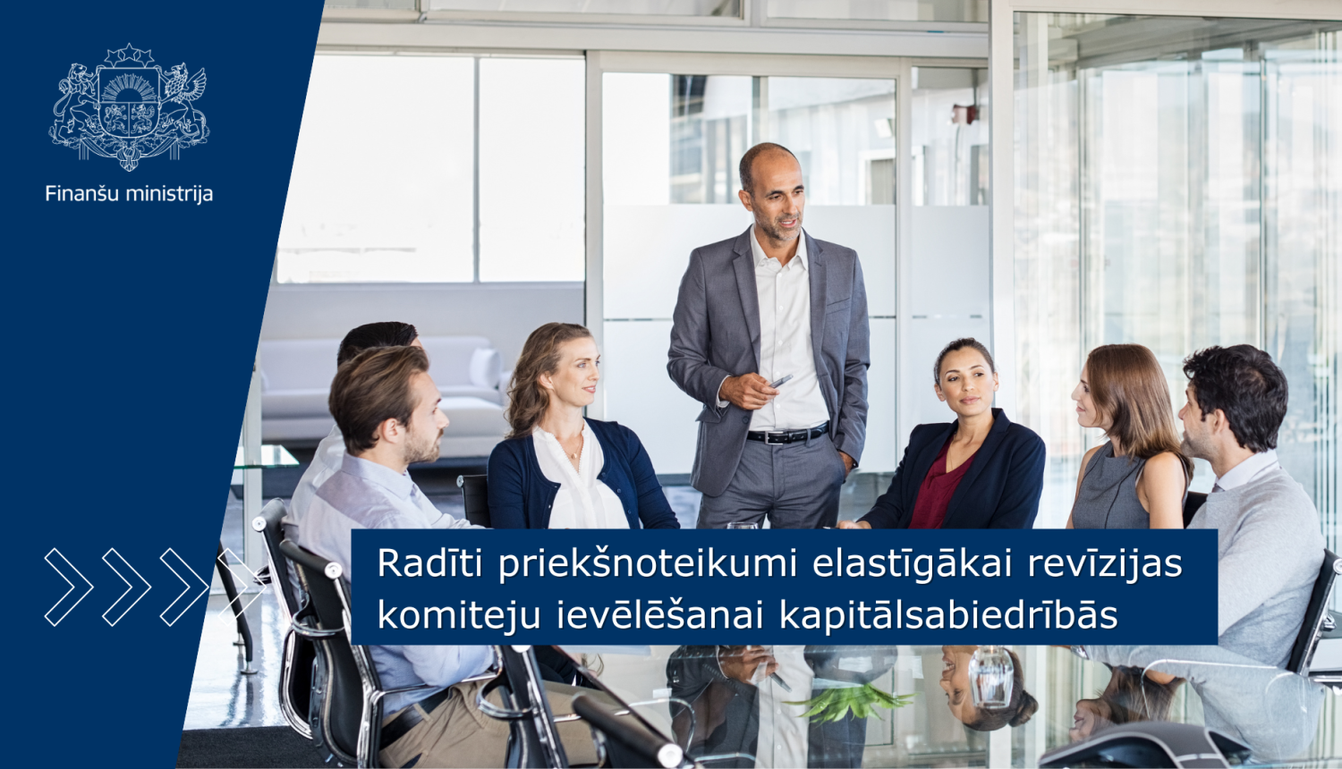 Birojā pie sanāksmju galda sēž sievietes un vīrieši, kas savstarpēji diskutē. viens vīrietis stāv pie galda