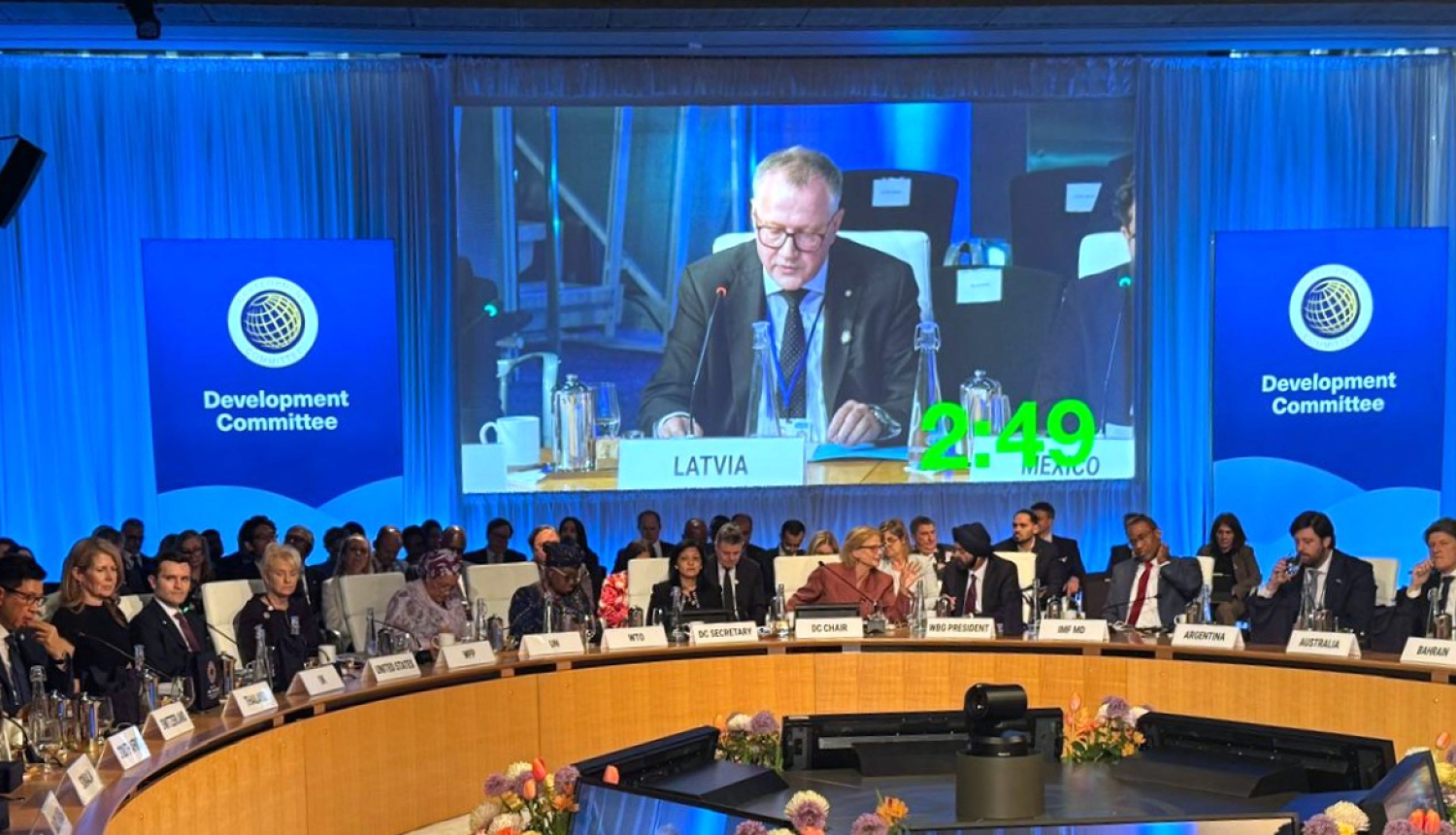 Konferenču zālē pie apaļa galda sēž delegāti no dažādām valstīm, ekrānā redzams finanšu ministrs uzstājoties ar runu