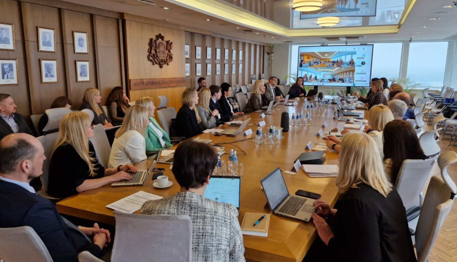 Pie gara galda sēž vīrieši un sievietes, savstarpēji sarunājas, uz galda stāv dokumenti un portatīvie datori, telpas galā ir redzams ekrāns uz kura tiek rādīta prezentācija