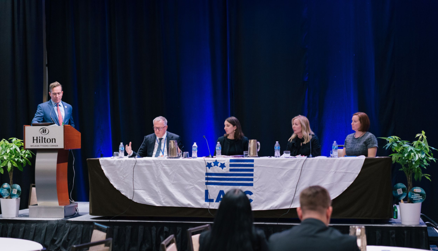 Attēlā pie tribīnes stāv vīrietis zilā uzvalkā, līdzās pie gara galda sēž četri cilvēki - sievietes un vīrieši - runā mikrofonos