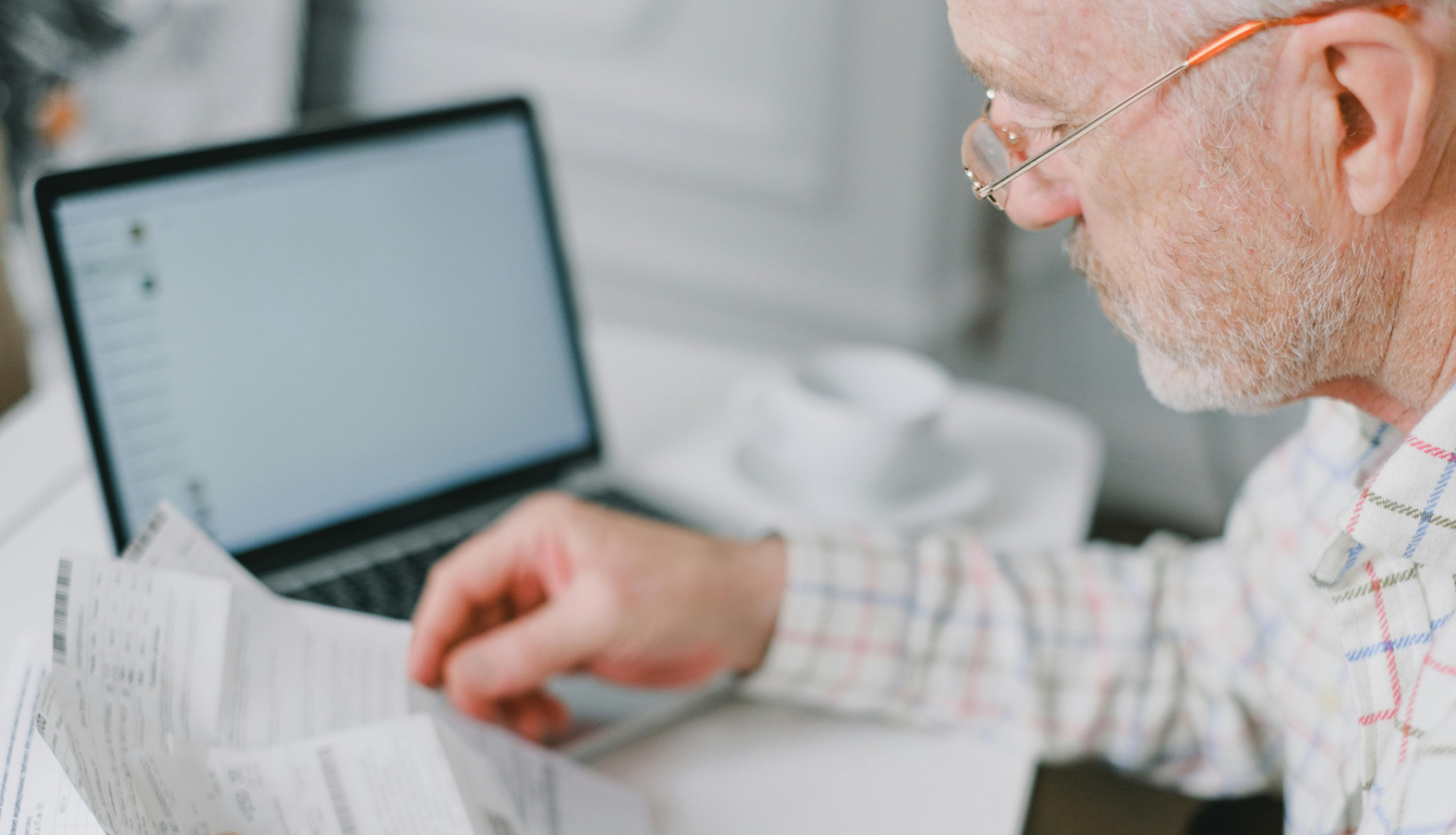 Persona ar brillēm pie darba galda pārskata papīra dokumentus, fonā atvērts klēpjdators