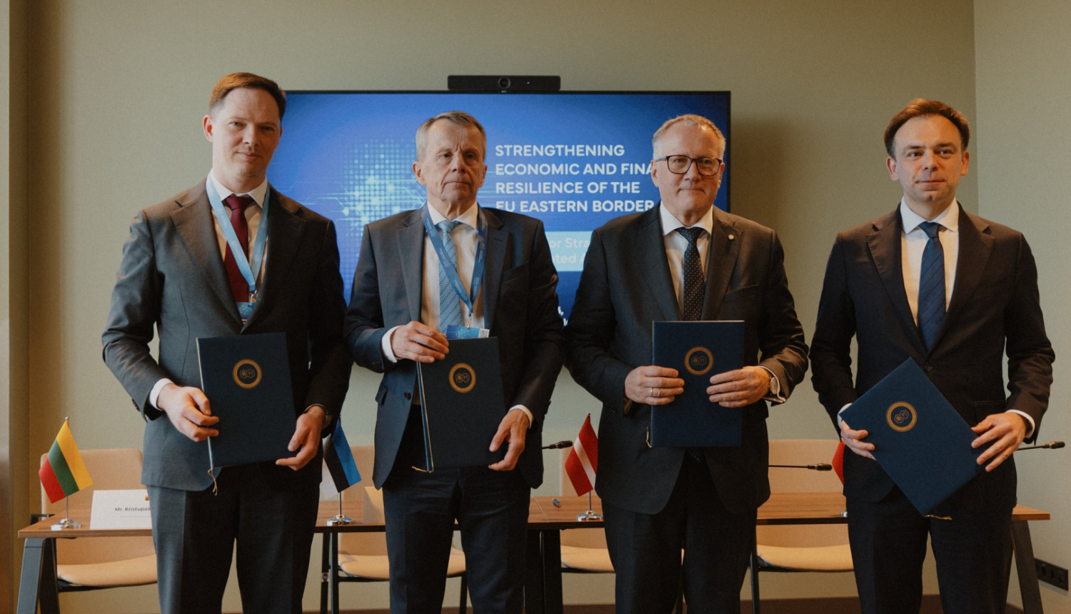 Četri vīrieši stāv rindā, tur rokās mapes ar dokumentiem, aiz viņu mugurām stāv gals ar krēsliem, pie sienas TV ekrāns