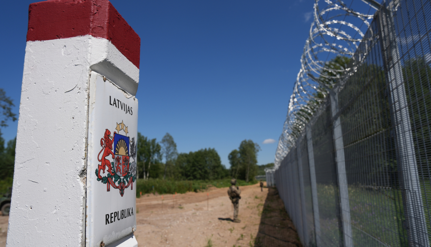 Attēlā tuvplānā redzams robežstabiņš ar Latvijas ģerboni, līdzās augstam žogam ar dzeloņstieplēm